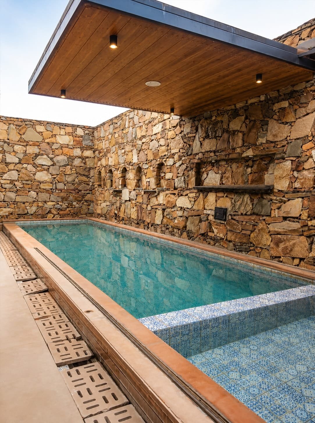 Pool with blue tiles under a wooden canopy against stone walls